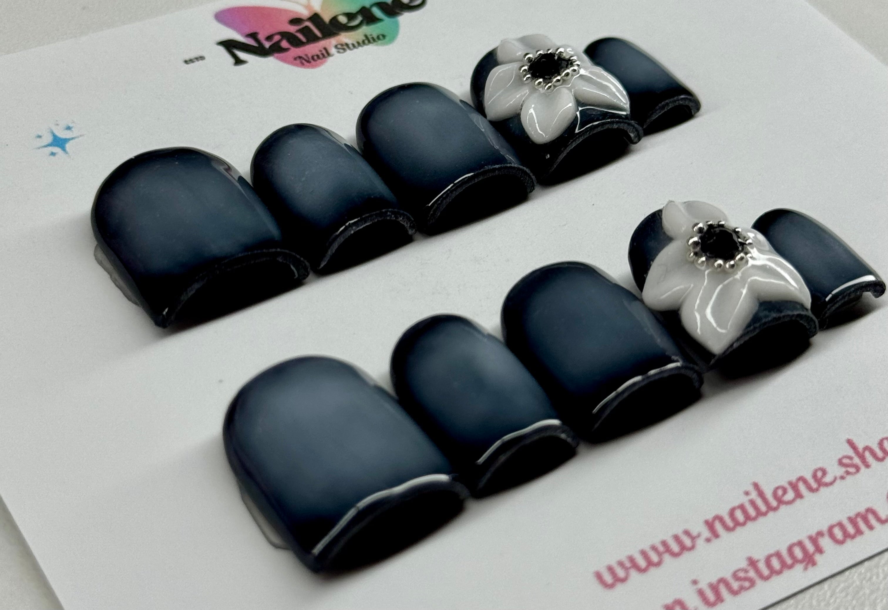 Set of black nail tips with white flower decorations on a branded card.